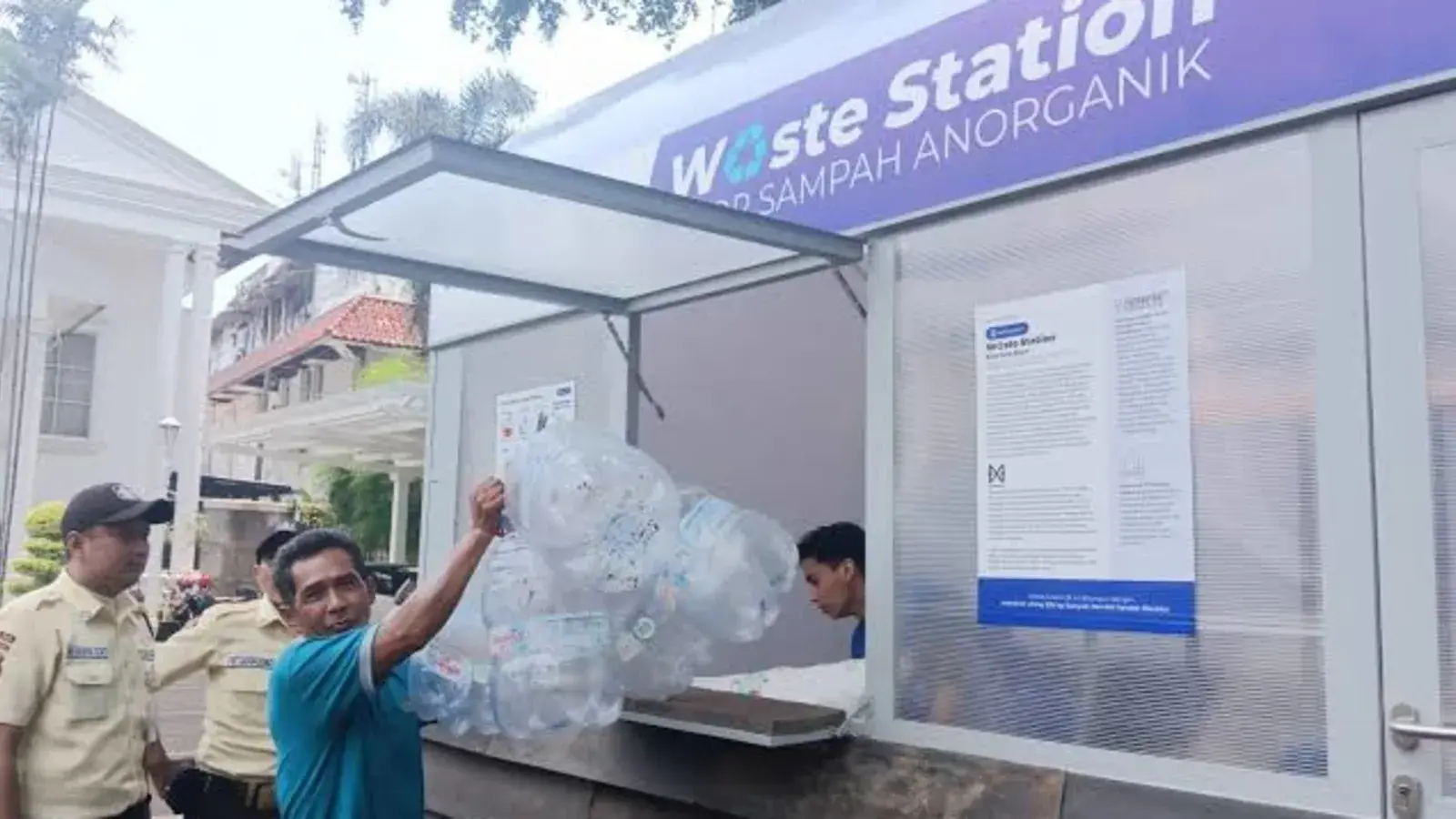 Residents handing sorted plastic bottles and bags of sachets to an attendant at a small waste drop-off kiosk in the grounds of Bogor City Hall, with a digital scale and a phone showing reward points