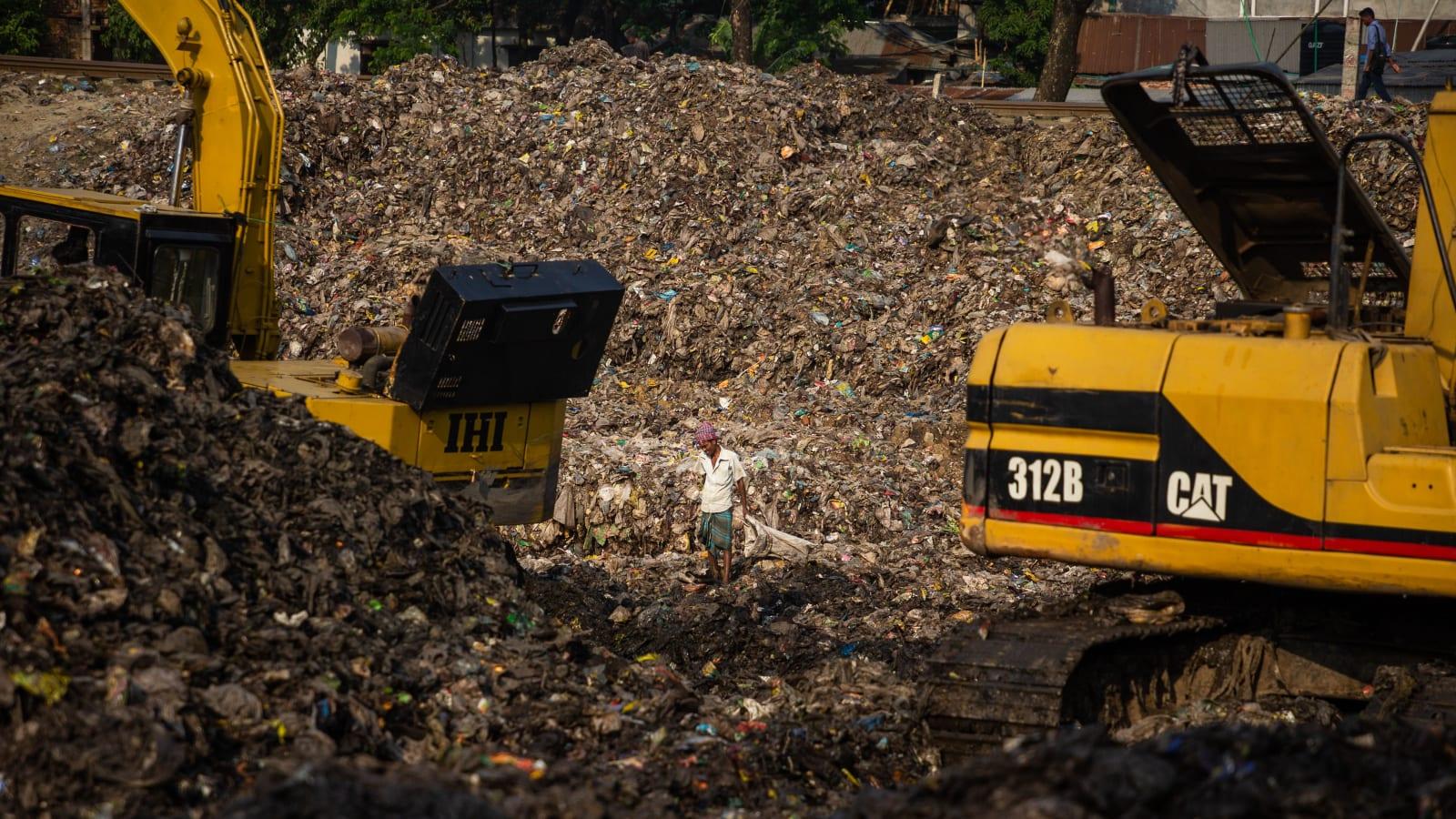 Refuse trucks queuing at a landfill working face where compacted waste is being covered with soil, with an excavator and cover material visible in the background